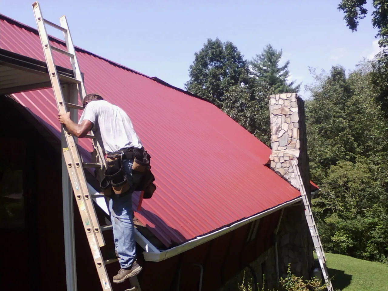 Roofer on a ladder beside a red metal roof and stone chimney — Soffit & Fascia Repair by Roofing Brothers in Decaturville